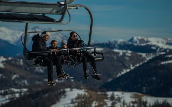 Family having fun on a ski lift with snow-covered mountains in the background during a sunny winter day.